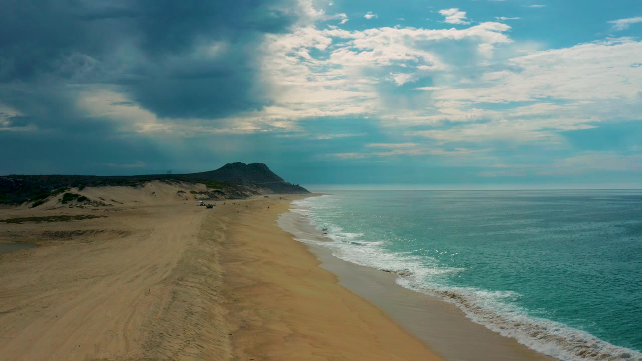 Aerial view of Laguna Beach in Baja California Sur, Mexico, with a long sandy shoreline, turquoise water, cloud-filtered sun, a distant headland, and no visible people.