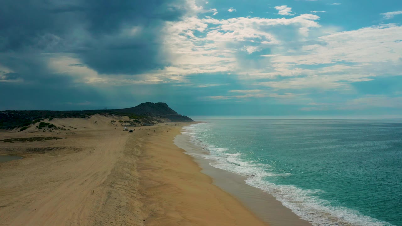 Aerial coastal view in Baja California Sur with turquoise water filling the right side, open sand on the left, a distant headland near the horizon, and no visible people.