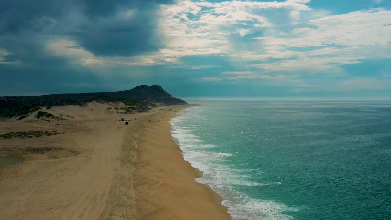 Aerial view one second into the clip showing Laguna Beach, Baja California Sur, with the shoreline angled farther into frame, pale surf edging the sand, dark hills inland, and no visible people.