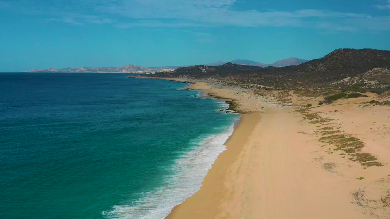 Aerial frame of Laguna Beach in Baja California Sur with the surf line bending along open sand, darker hills behind the beach, distant land on the horizon, and no visible people.
