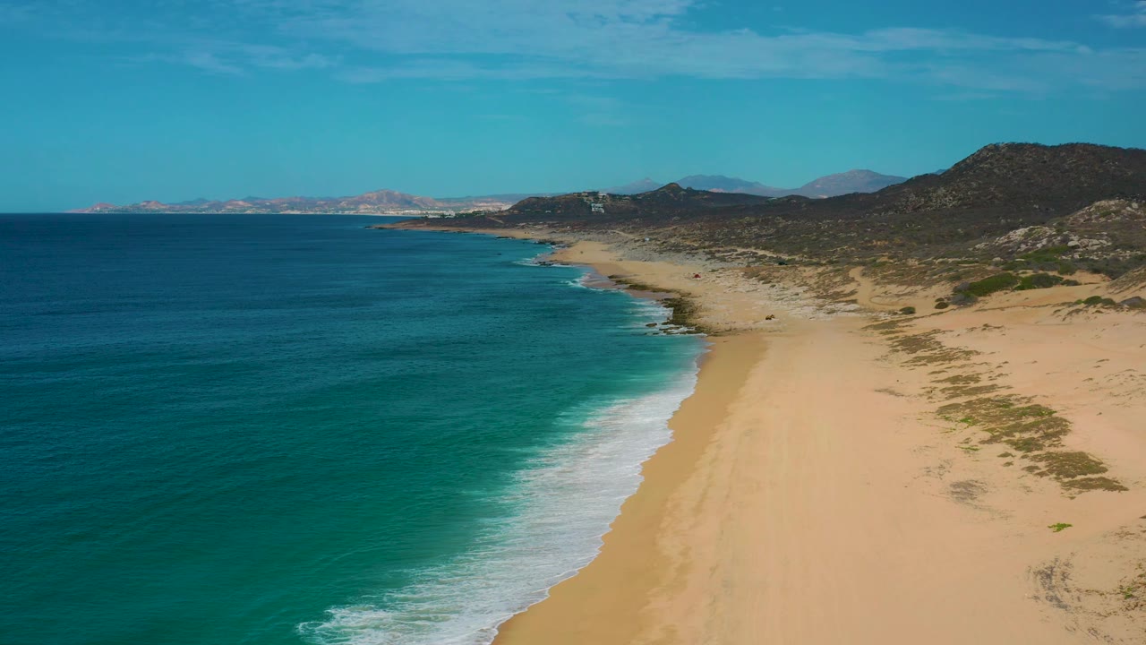 Drone view over Laguna Beach, Baja California Sur, looking along the sandy shore as white waves meet the beach, scrub patches spread across the dunes, and no visible people appear.