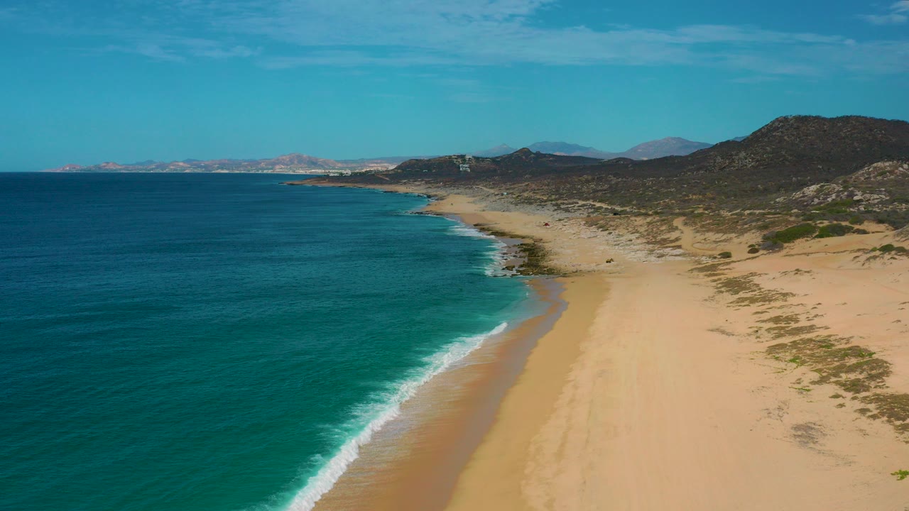 Aerial view over Laguna Beach, Baja California Sur, with deep blue ocean on the left, pale sand on the right, low hills inland, and no visible people.