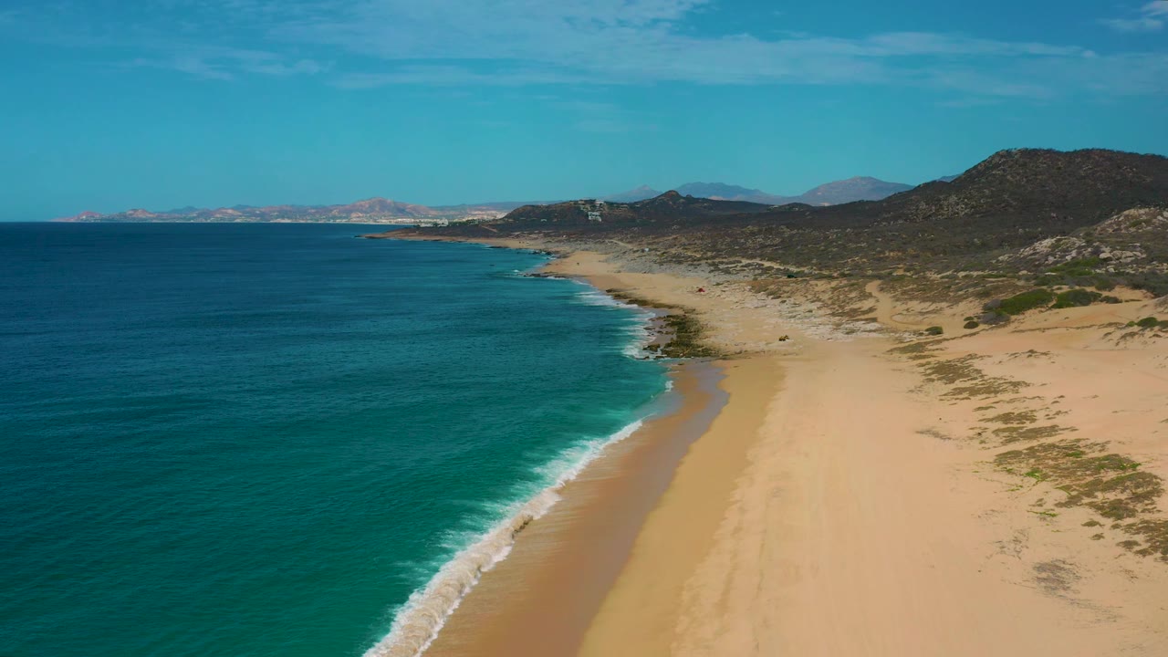 Wide aerial frame at Laguna Beach, Baja California Sur, with turquoise ocean water pushing a white wave edge onto the sand and no visible people on the beach.