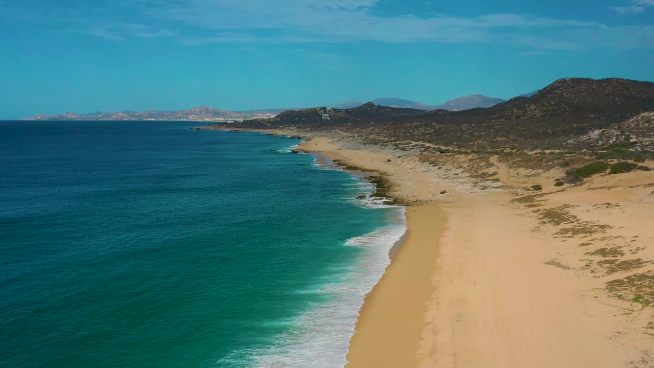 Aerial frame over Laguna Beach, Baja California Sur, showing the surf line closer to center, wide sand to the right, low scrub, dark hills, blue sky, and no visible people.
