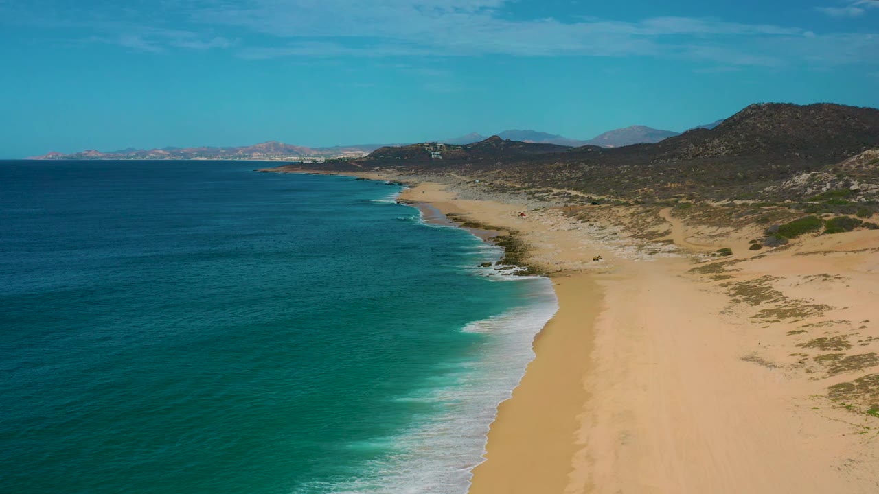 High aerial view of Laguna Beach, Baja California Sur, with turquoise water filling the left side, white wash along the beach edge, dune scrub to the right, distant hills, and no visible people.