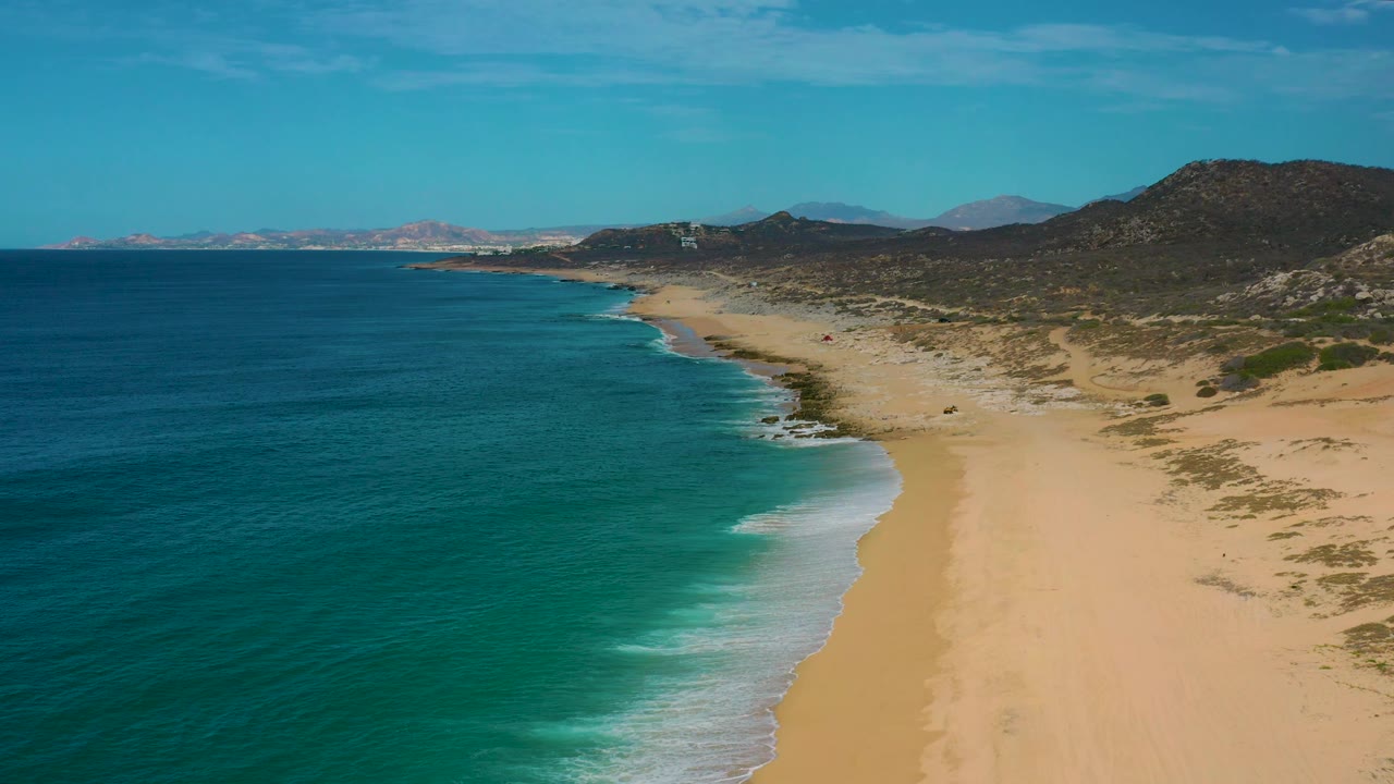 Mid-clip aerial view of Laguna Beach, Baja California Sur, looking along a quiet beach where the surf pushes diagonally across the sand, with hills beyond and no visible people.