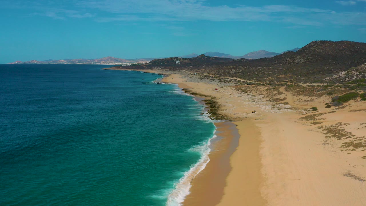 Aerial view over Laguna Beach, Baja California Sur, with turquoise water left of a pale sandy shoreline, low mountains in the background, and no visible people.