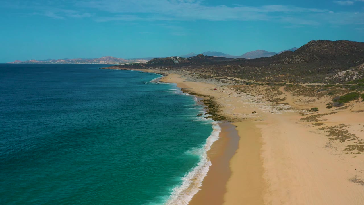 Wide aerial frame over Laguna Beach, Baja California Sur, with open sand in the foreground, white surf tracing the waterline, low mountains to the right, and no visible people.