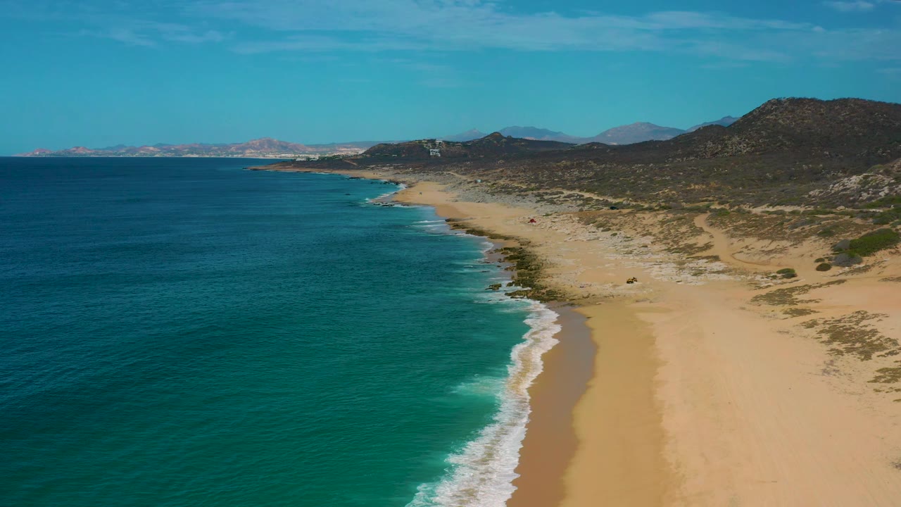 Aerial coastline frame from Laguna Beach, Baja California Sur, with teal ocean on the left, a narrow band of breaking white waves, scrub-covered dunes, rocky low hills, and no visible people.