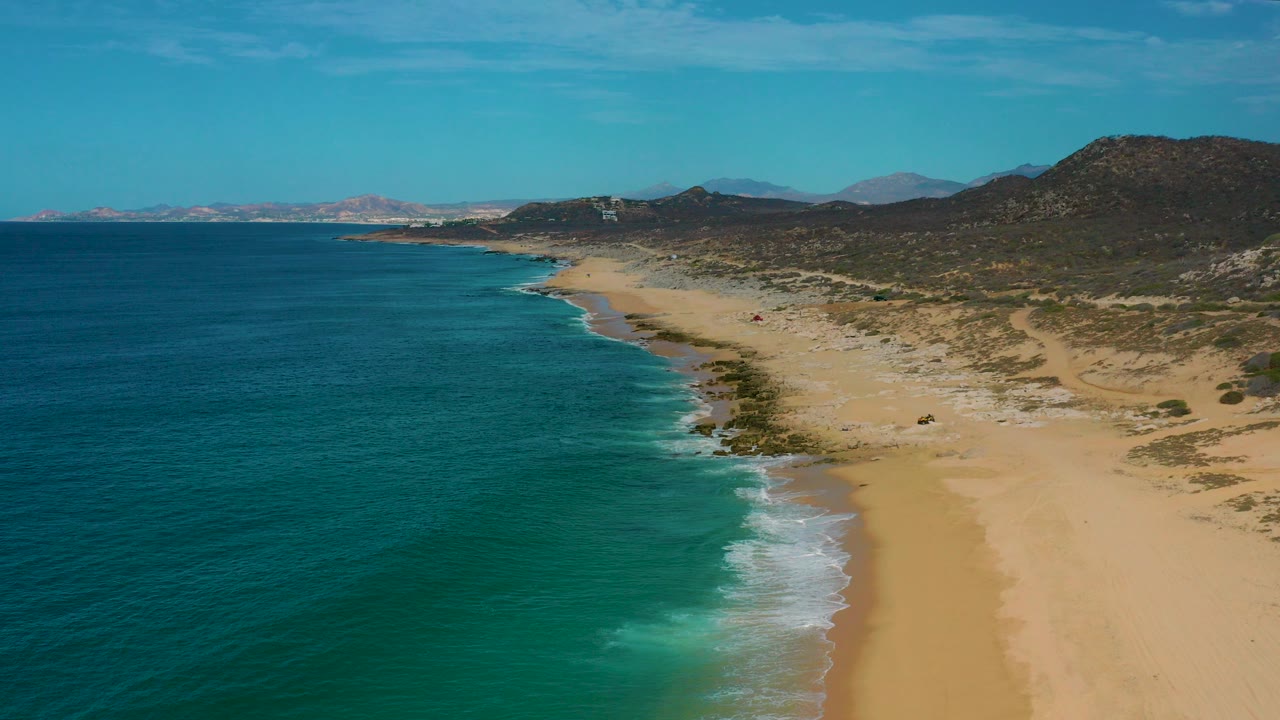 Opening aerial frame over Laguna Beach, Baja California Sur, with turquoise water on the left, pale sand on the right, low hills in the distance, and no visible people.
