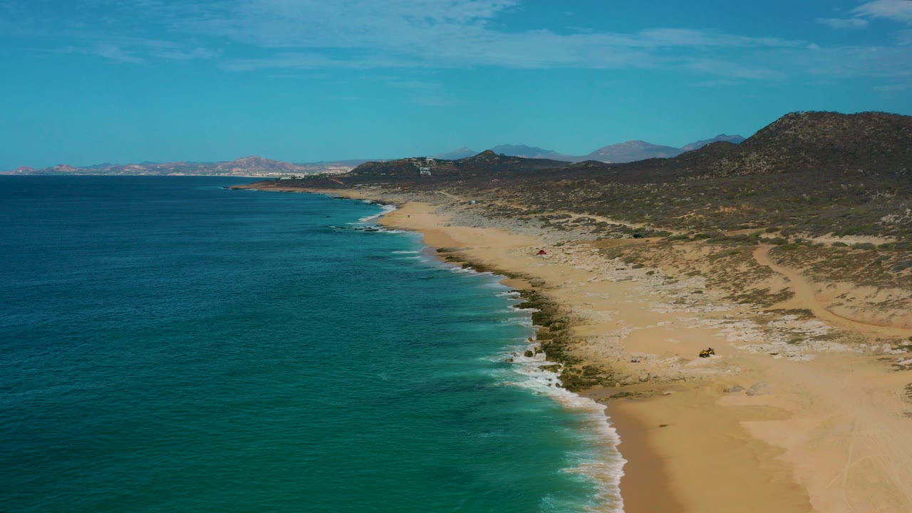 Aerial view of Laguna Beach, Baja California Sur with blue ocean on the left, a pale sandy beach curving along the shore, rocky hills to the right, and no visible people.