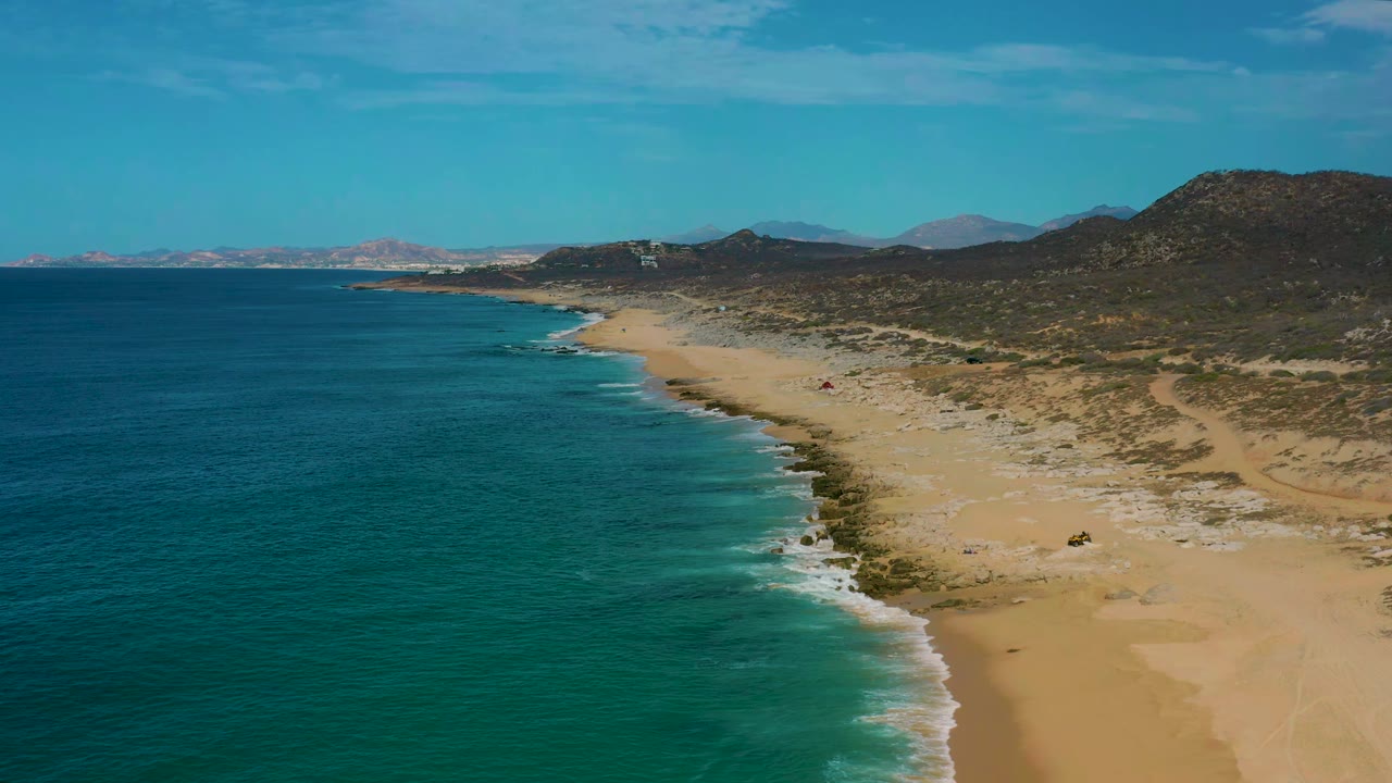 Aerial frame over Laguna Beach, Baja California Sur showing turquoise water grading darker offshore, a long sand strip, dry hillside terrain, and no visible people.