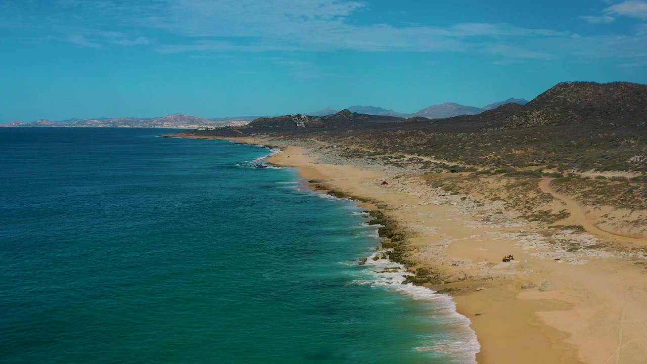 Aerial view at Laguna Beach, Baja California Sur with the beach running diagonally through frame, ocean filling the left side, mountain terrain beyond the sand, and no visible people.