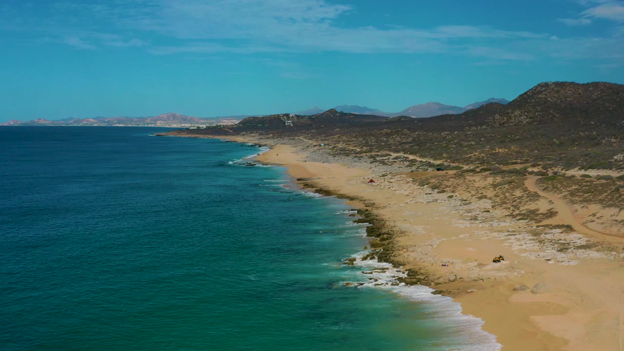 Aerial frame from Laguna Beach, Baja California Sur showing small white waves reaching a broad sand beach, dark blue open water to the left, scrubby slopes to the right, and no visible people.