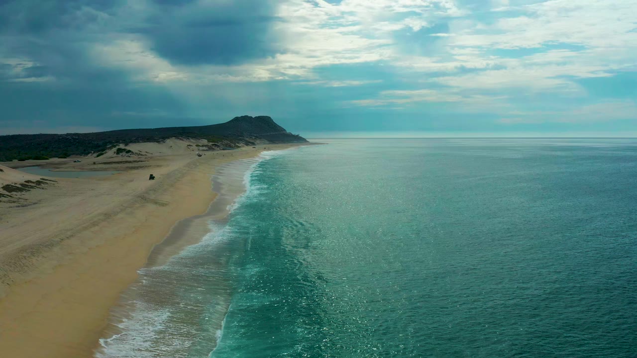 Aerial frame of Laguna Beach, Baja California Sur, Mexico, with white surf tracing a sandy shore, open turquoise water on the right, a distant headland ahead, cloud cover above, and no visible people.