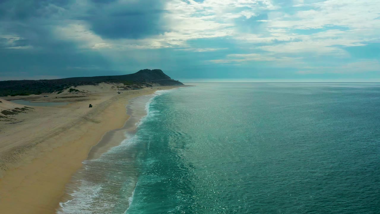 Aerial frame of Laguna Beach, Baja California Sur, Mexico, showing a long sand strip on the left, a curved band of breaking waves near center, broad ocean on the right, heavy clouds overhead, and no visible people.