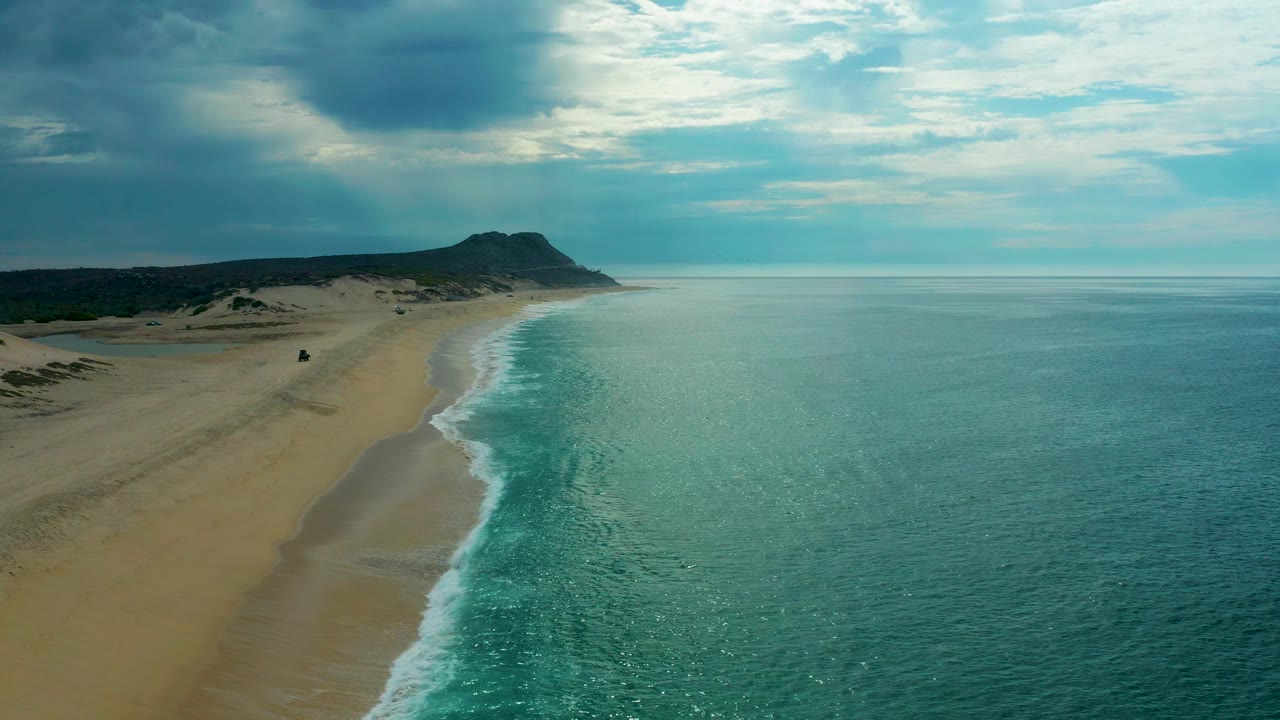 Aerial frame of Laguna Beach, Baja California Sur, Mexico, where the sandy beach occupies more of the left side, the surf breaks in a bright white strip near center, the ocean lies calm to the right, and no visible people appear.