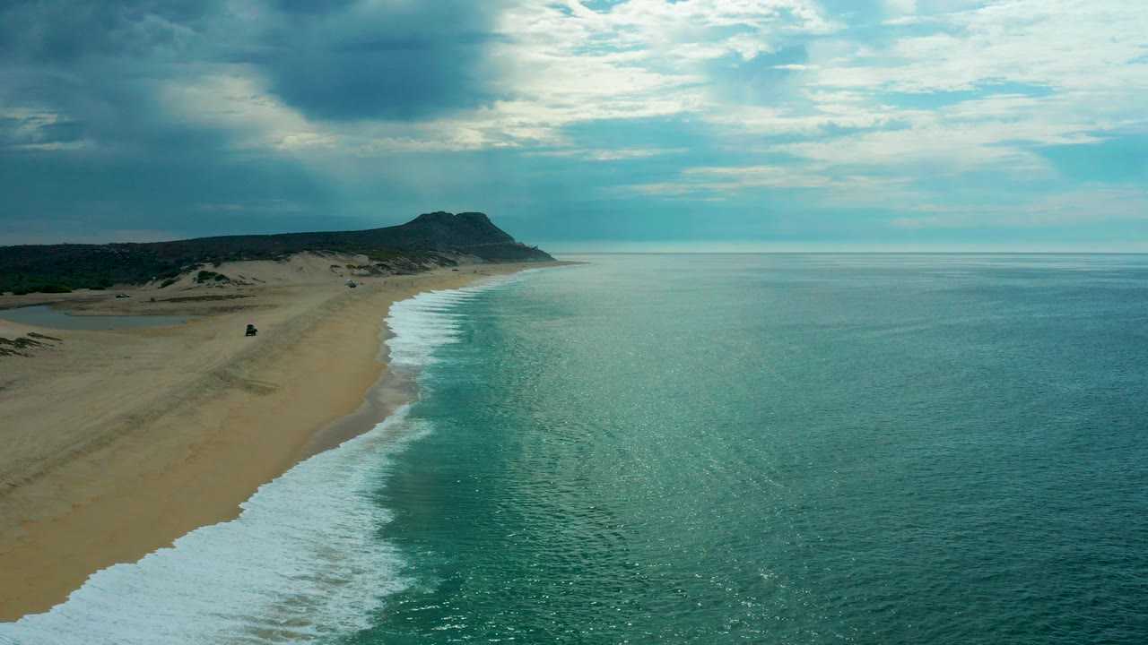 Aerial view of Laguna Beach in Baja California Sur with sand along the left edge, turquoise water on the right, a distant headland ahead, and no visible people.