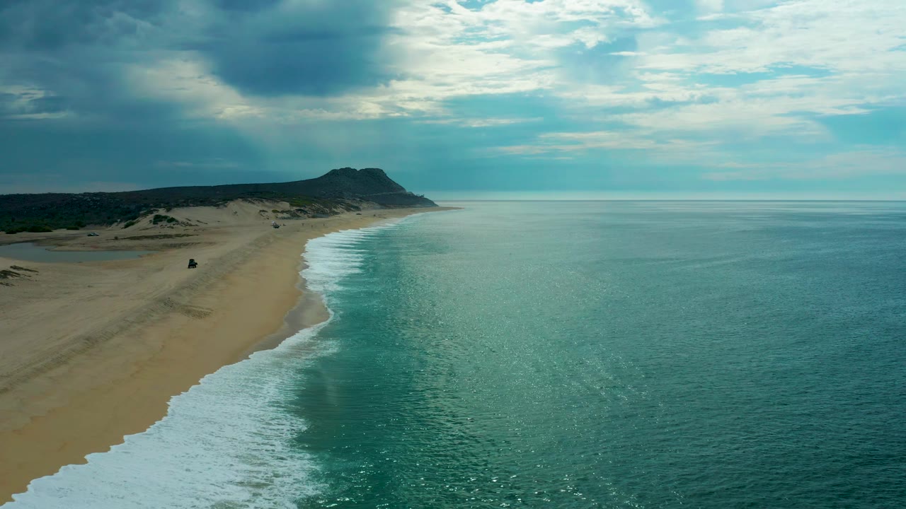 Wide aerial view of Laguna Beach in Baja California Sur with more open ocean to the right, pale surf tracing the sand, dark cloud patches above, and no visible people.