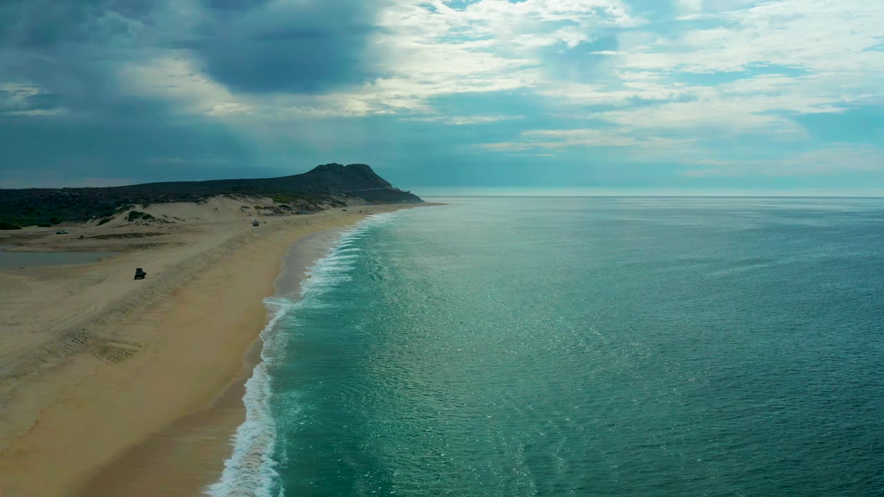 Laguna Beach coastline in Baja California Sur seen from above, with the surf line slightly farther down frame, open sand to the left, calm teal water to the right, and no visible people.