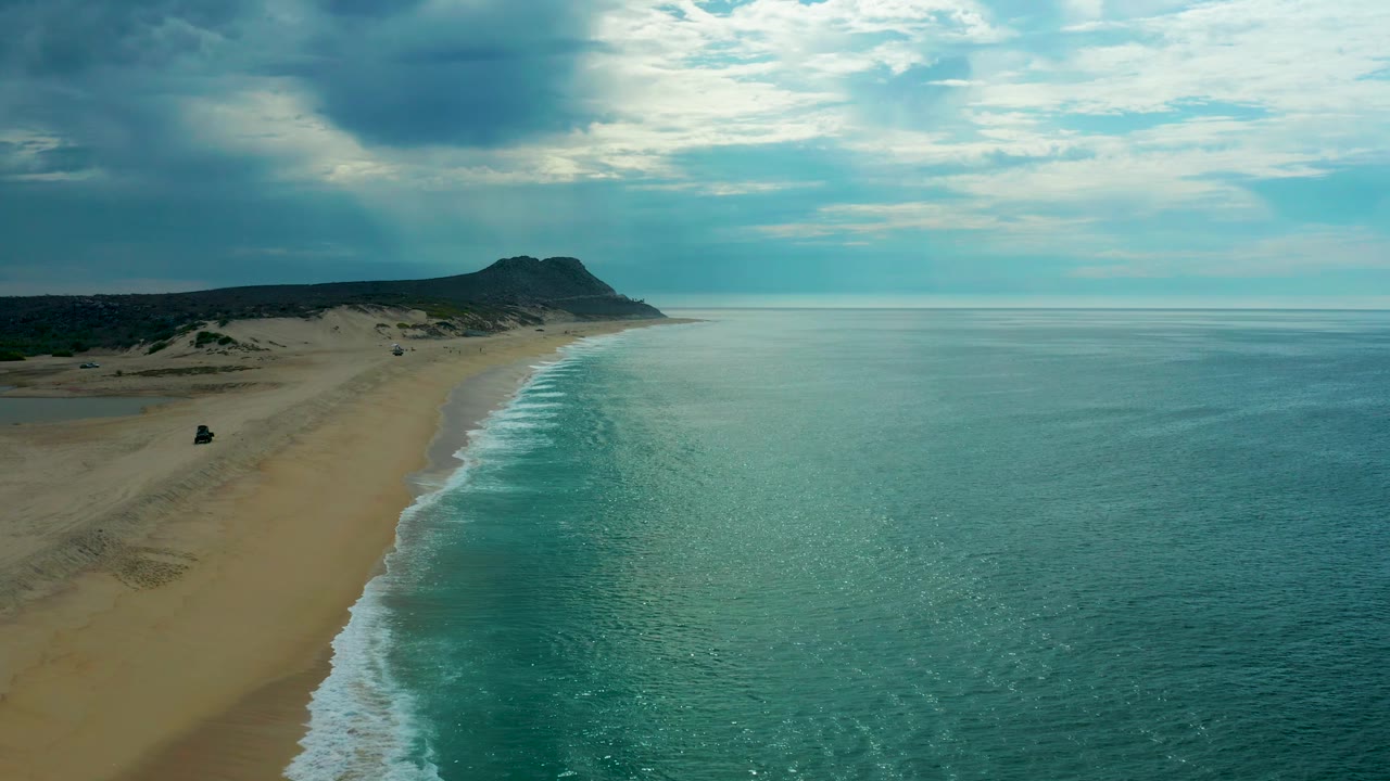 A wide aerial view of Laguna Beach, Baja California Sur, with the ocean occupying most of the right side, a pale sandy beach along the left, low hills beyond, cloud-filtered light, and no visible people.