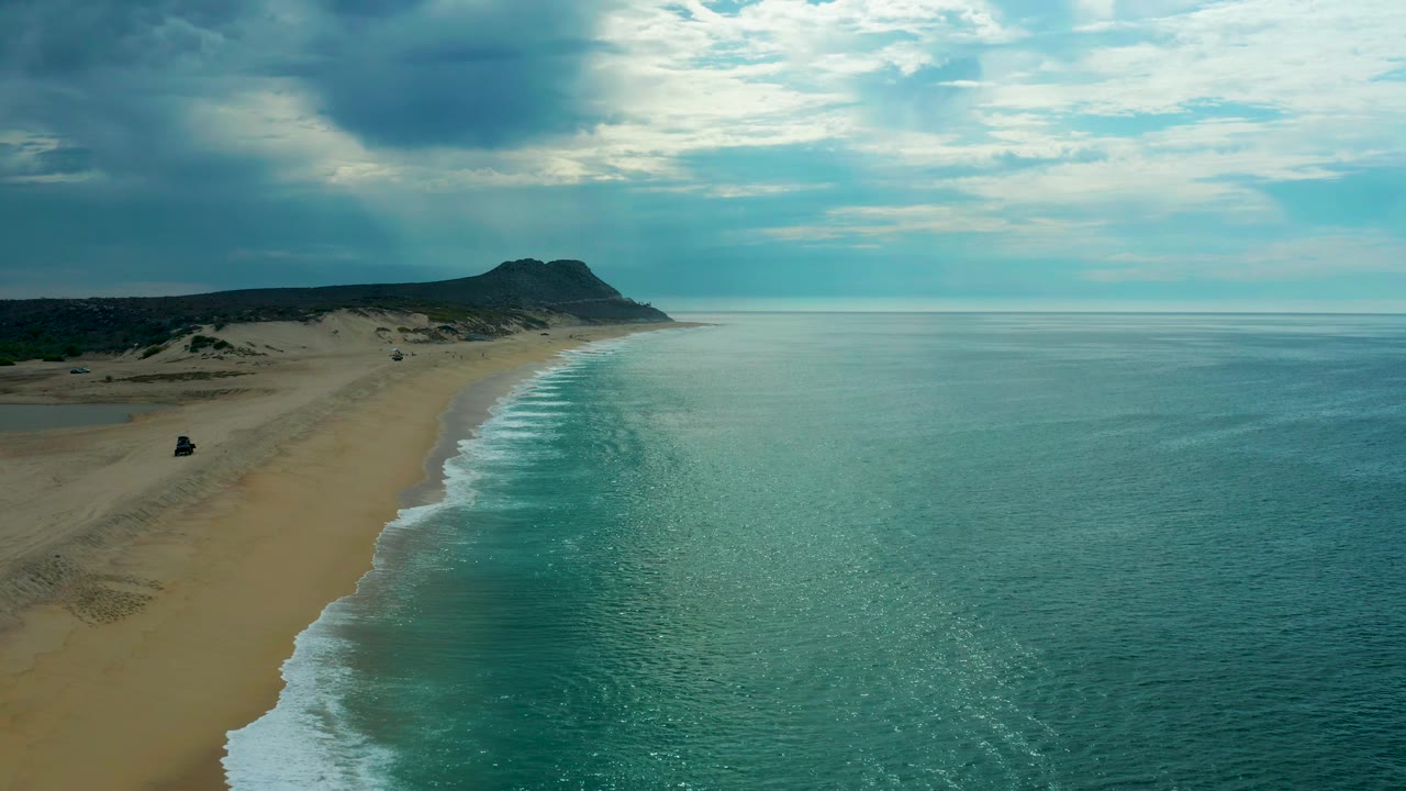 Laguna Beach in Baja California Sur appears from an elevated coastal angle, with empty sand stretching toward a distant headland, mild surf marking the shoreline, blue-gray clouds overhead, and no visible people.
