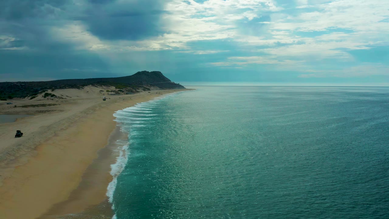Aerial view along Laguna Beach, Baja California Sur, with the surf line running diagonally, low dunes behind the sand, open ocean to the right, and no visible people.