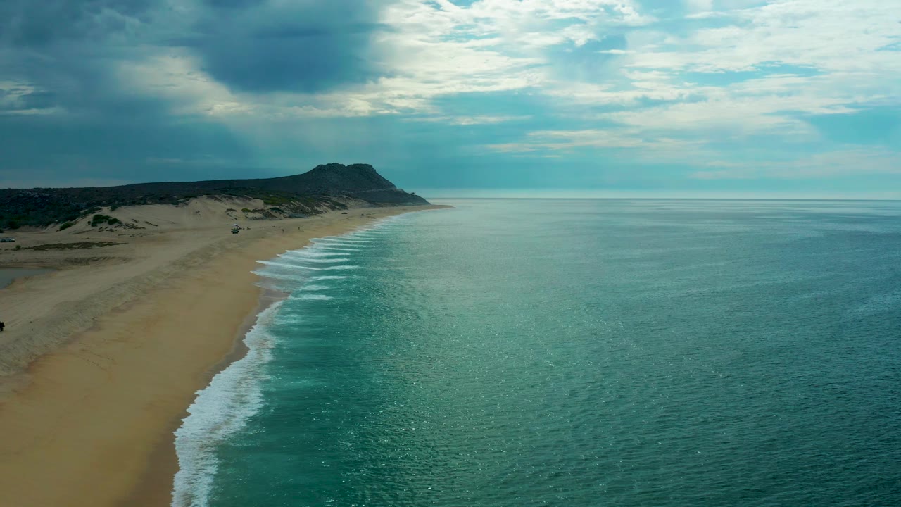Mid-clip aerial frame over Laguna Beach, Baja California Sur, showing more open ocean across the right side, the sandy shore along the left, layered clouds above, and no visible people.