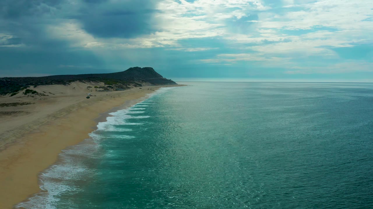 Laguna Beach coastline from above, with more open water filling the right side, surf tracing the sand, a low headland ahead, and no visible people.