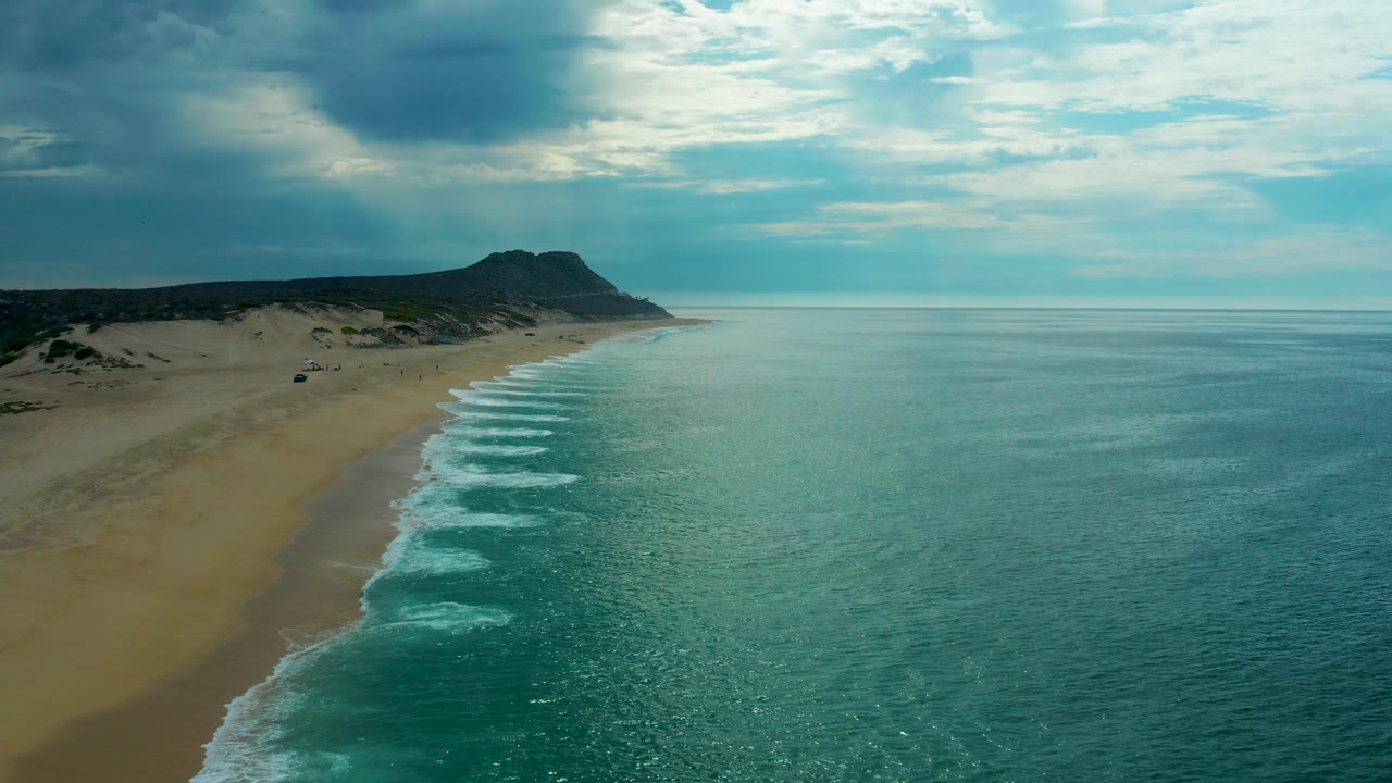 Aerial view of a sandy beach beside turquoise ocean under clouds.