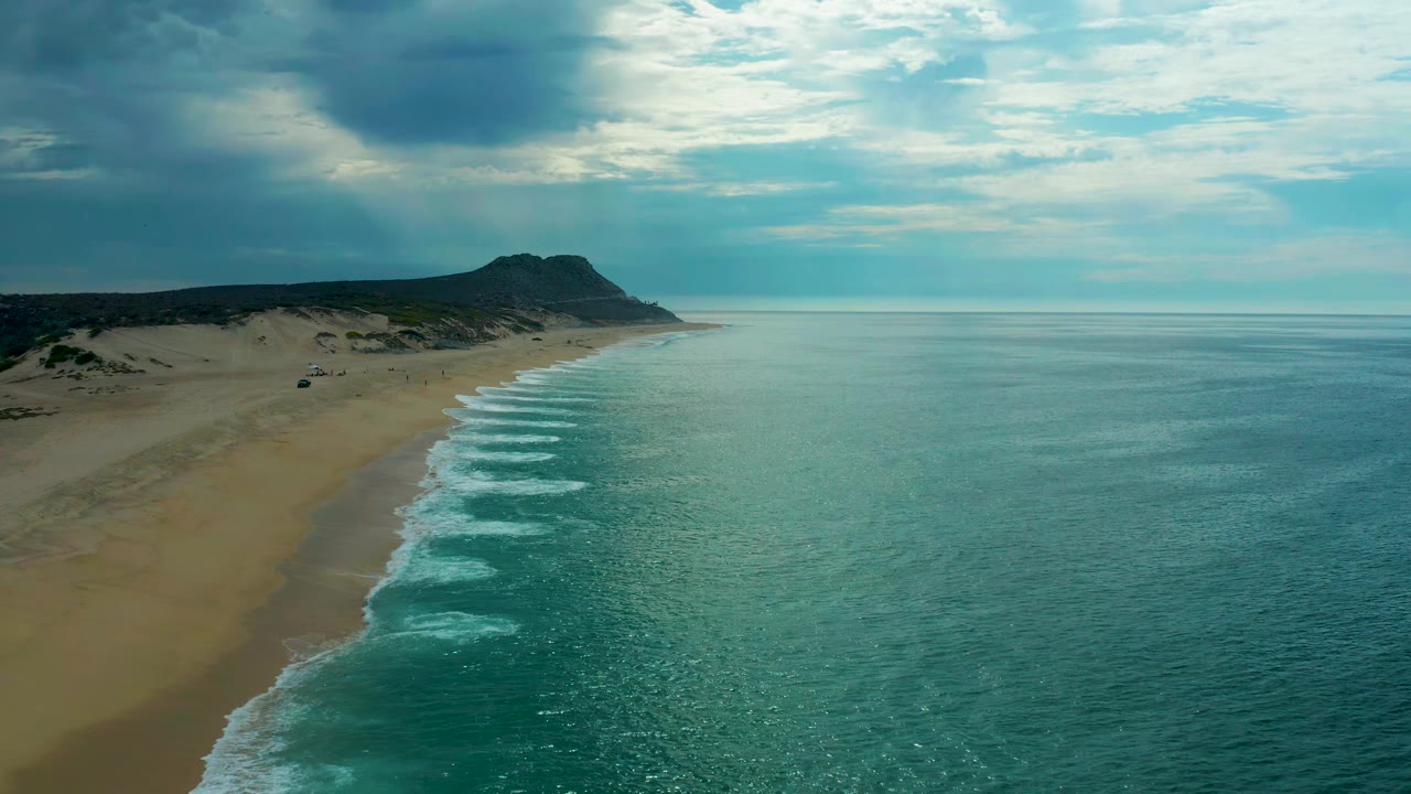 Aerial frame over Laguna Beach, Baja California Sur, looking along the shoreline as white wave bands meet the sand, with open ocean to the right and no visible people.