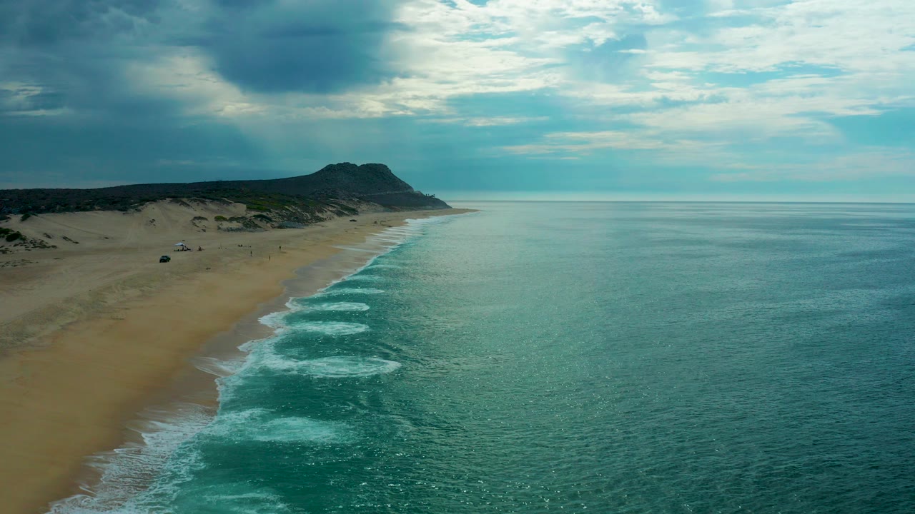 Aerial view of Laguna Beach in Baja California Sur with surf bands reaching a sandy shoreline, a dark headland in the distance, heavy clouds overhead, and no visible people.