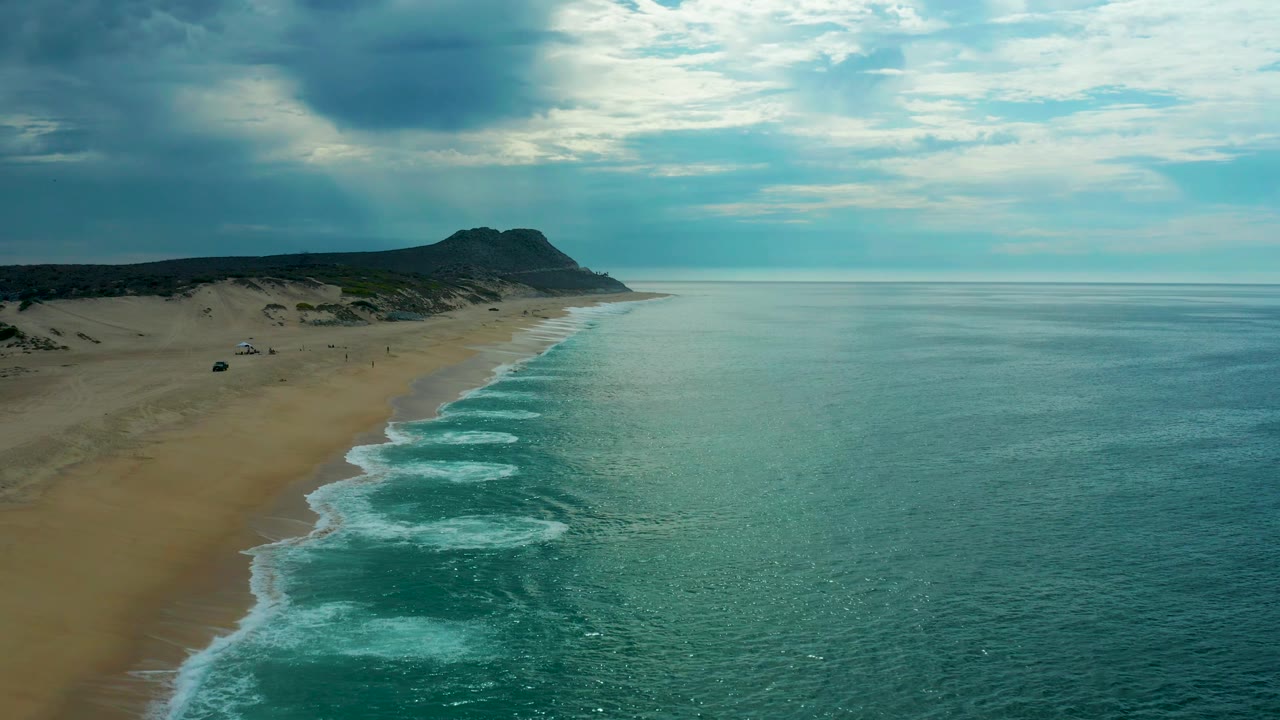 Wide aerial frame over Laguna Beach, Baja California Sur, showing a long sandy coast, multiple white surf lines, smooth open ocean, heavy cloud cover, and no visible people.