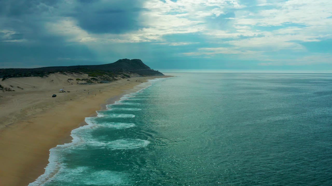 Aerial view along Laguna Beach with the sandy strip closer in frame, pale surf rolling in rows, a dark cloud mass overhead, and no visible people.
