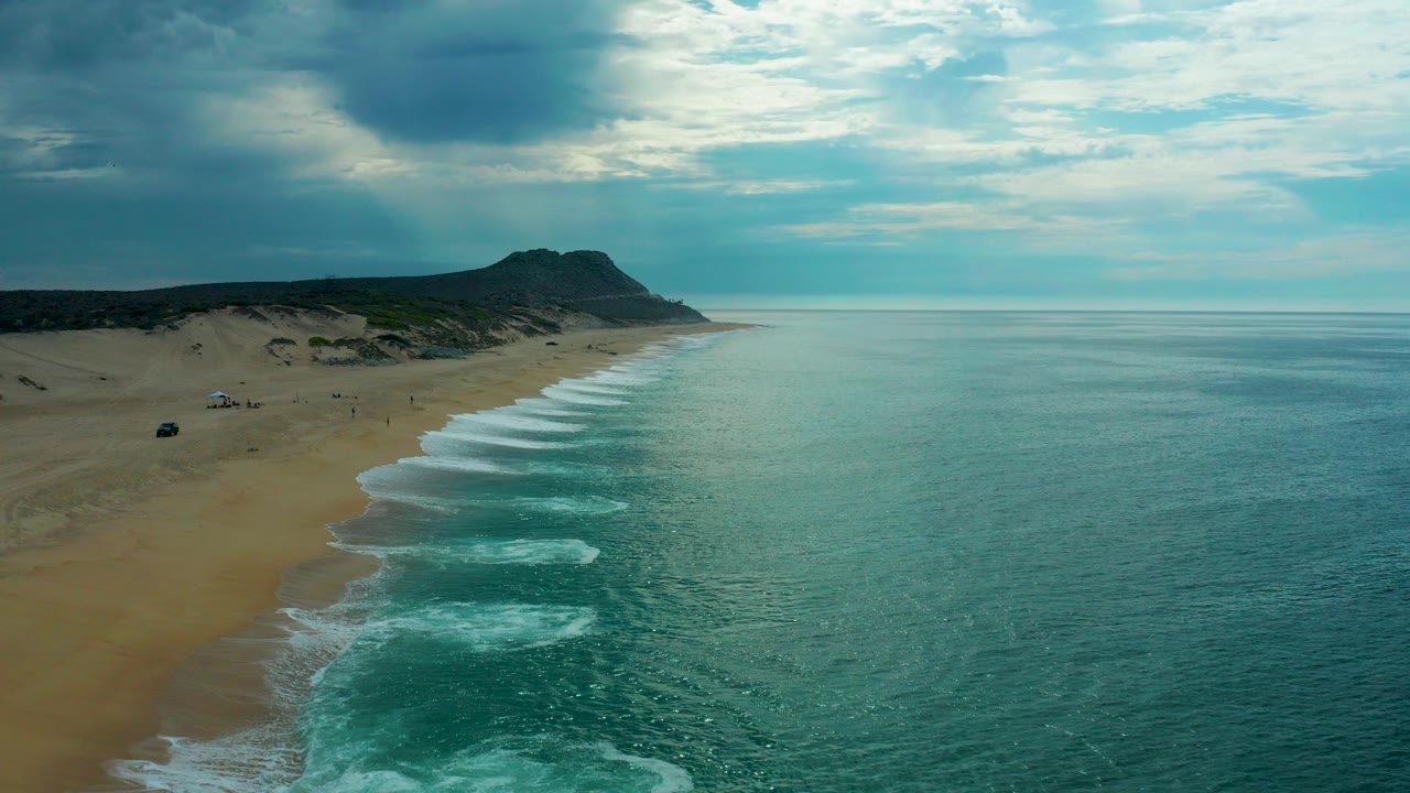 Laguna Beach, Baja California Sur aerial frame with sandy shoreline on the left, turquoise ocean on the right, white surf bands, and a dark headland under broken clouds.
