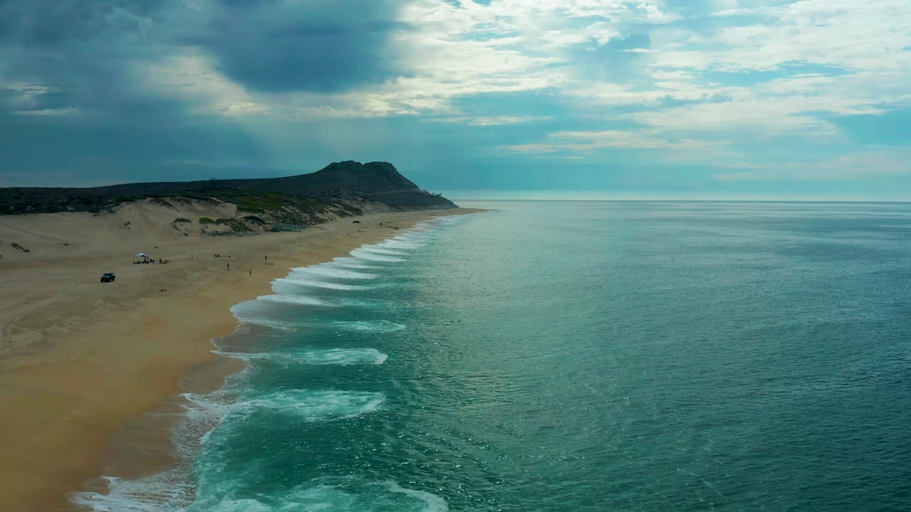 Laguna Beach, Baja California Sur coastline seen from above, with the beach running diagonally toward a low headland and foamy waves spreading across teal water.