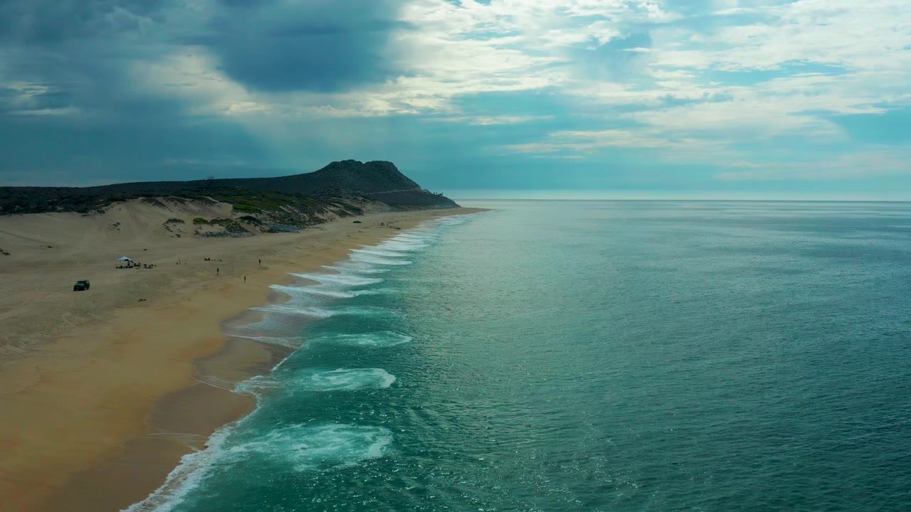 Laguna Beach, Baja California Sur beach from above, with small beach vehicles or setups on the sand, repeating surf arcs, teal water, and a dark cloud bank overhead.