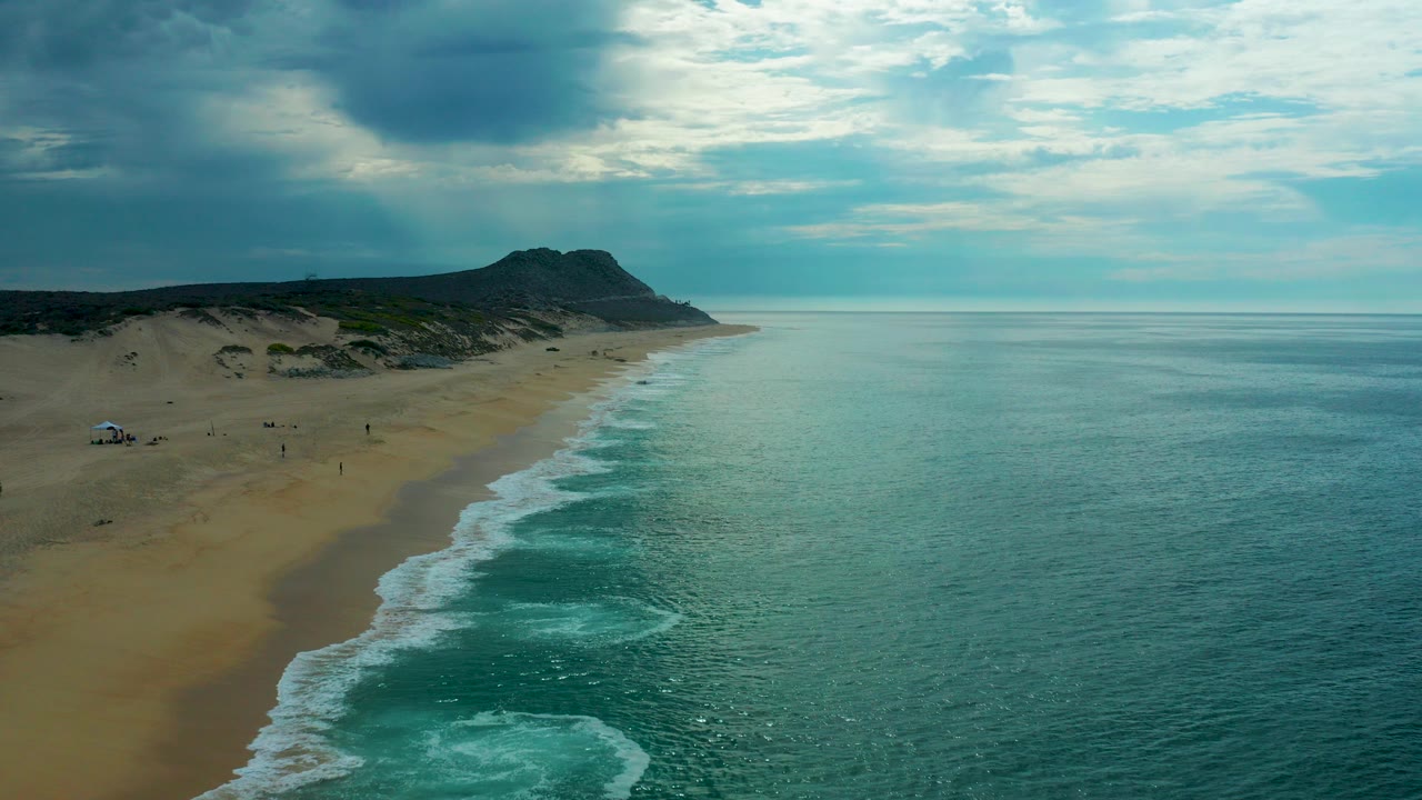 Aerial frame over Laguna Beach, Baja California Sur with foamy surf crossing a sandy shoreline, dunes and a dark headland in the background, and no recognizable people visible.