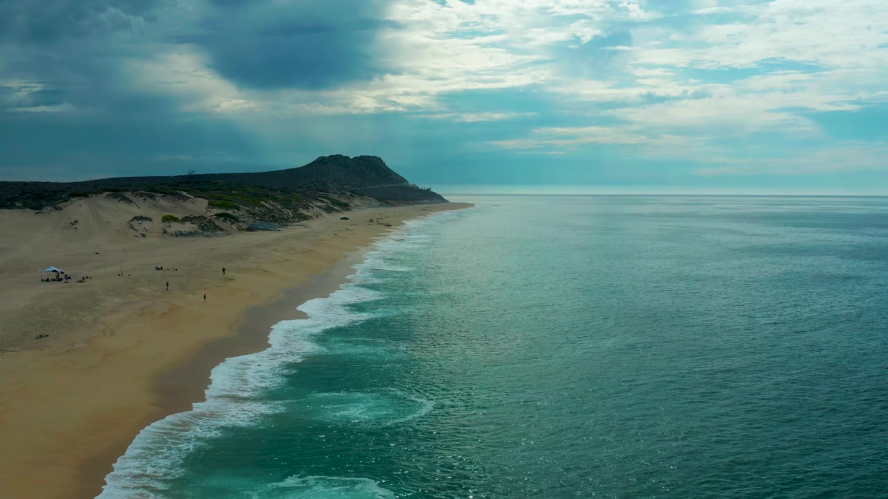 Aerial view at Laguna Beach, Baja California Sur where a fresh band of foam curls onto the sand, the sea fills most of the right frame, and muted clouds cover the sky.