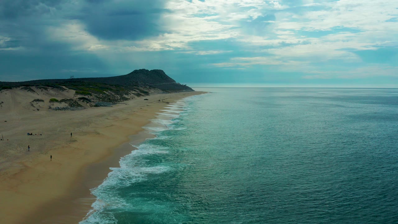 Opening aerial frame over Laguna Beach, Baja California Sur, with a diagonal sandy shoreline, teal sea, white surf bands, dark dune ridge, cloudy sky, and tiny distant figures on the beach.