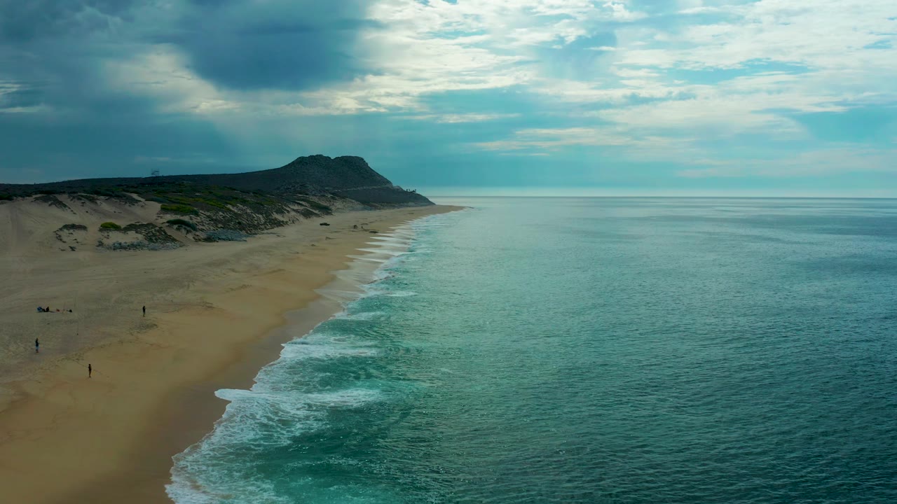 Aerial frame over Laguna Beach, Baja California Sur, with the long beach receding toward a dark headland, calm teal water filling the right side, low clouds above, and small distant figures near the sand.