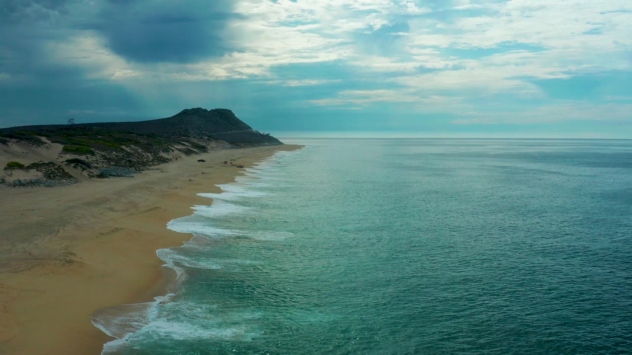 Aerial view at Laguna Beach, Baja California Sur, with turquoise water meeting a wide sandy shore, dark dunes and a distant headland under clouded light, with no visible people.
