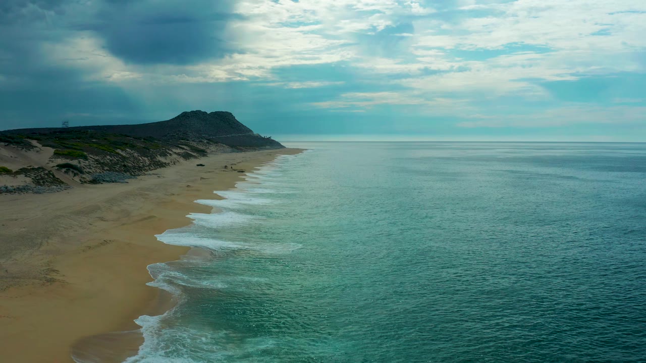 Wide aerial view of Laguna Beach in Baja California Sur with the shoreline centered between teal ocean and empty sand, dark dunes behind the beach, and no visible people.