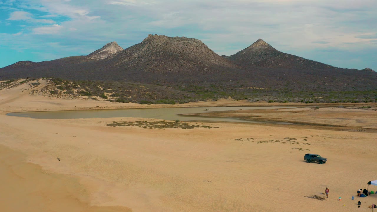 Wide aerial frame over Laguna Beach, Baja California Sur, with pale sand in front, a lagoon channel, rocky mountains, a dark jeep, and no recognizable people visible.