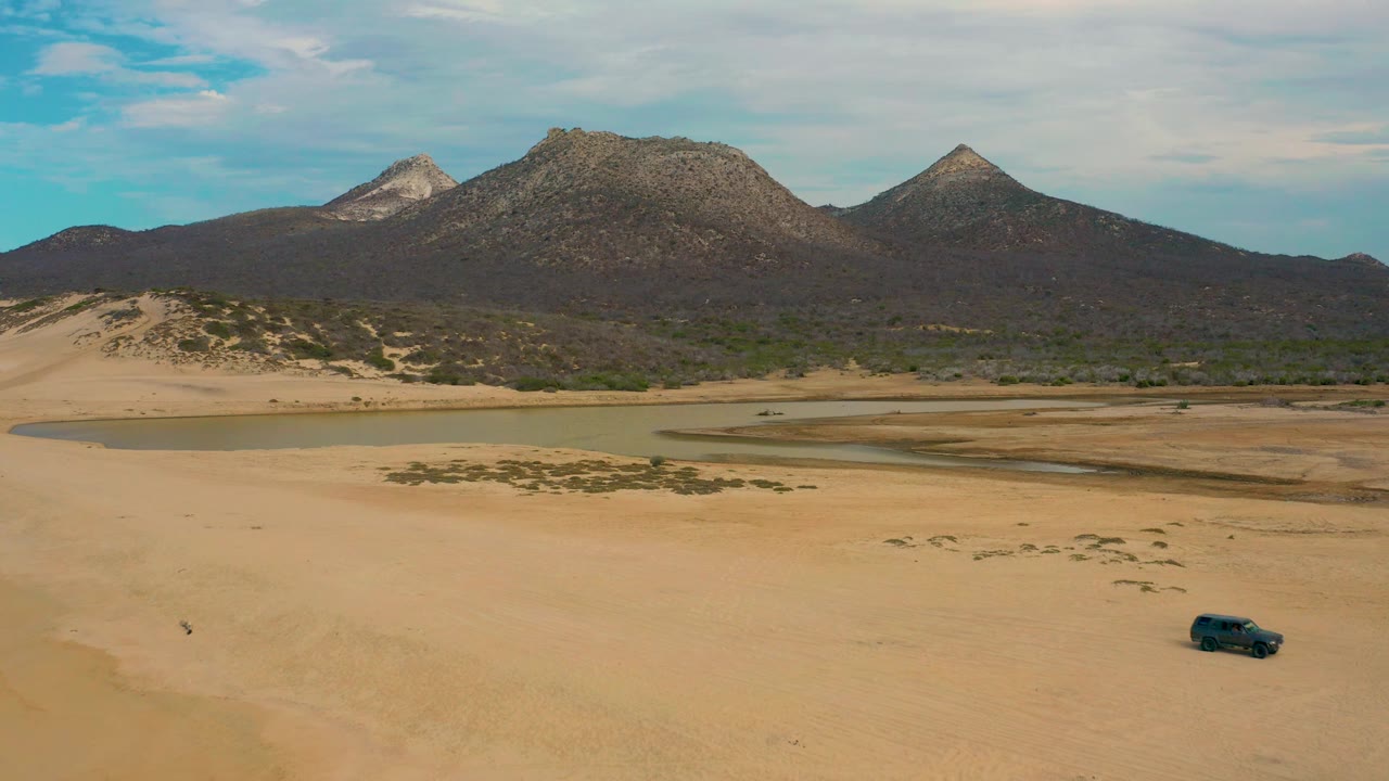 High view over Laguna Beach, Baja California Sur, with the jeep near the right side of the sandy foreground, lagoon water across the middle, and three rocky peaks in the distance.