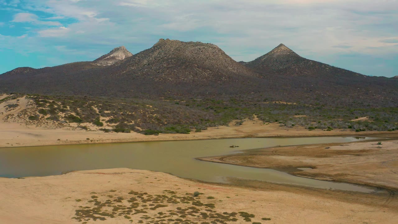 Aerial view at Laguna Beach in Baja California Sur showing a curved lagoon edge, sandy flats, scattered desert brush, dark mountain slopes, blue sky, white clouds, and no visible people.