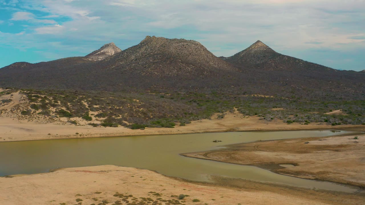 Laguna Beach, Baja California Sur aerial with lagoon water across the lower frame, a dry shoreline, scrub-covered hills, three rocky peaks, blue sky, light clouds, and no visible people.