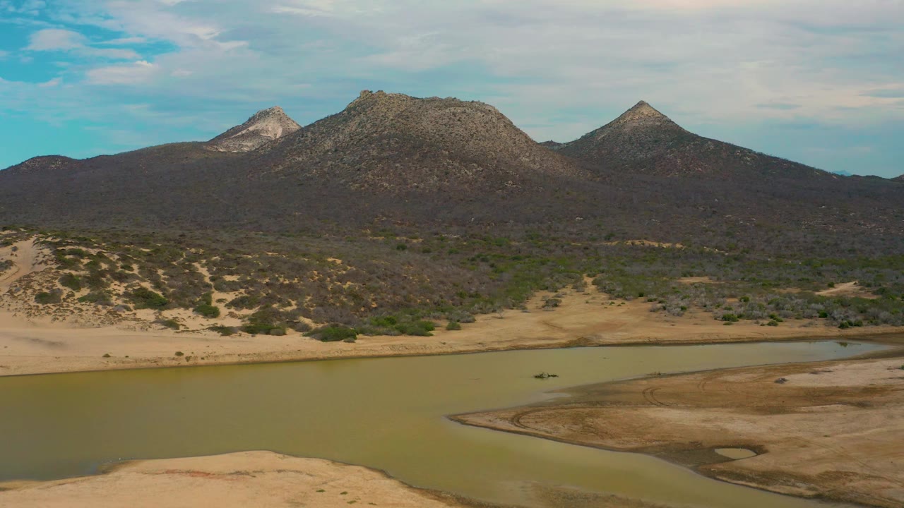 Drone-height view in Laguna Beach, Baja California Sur with green lagoon water and a sandbar in the foreground, scrub terrain beyond it, rocky mountains, cloudy blue sky, and no visible people.