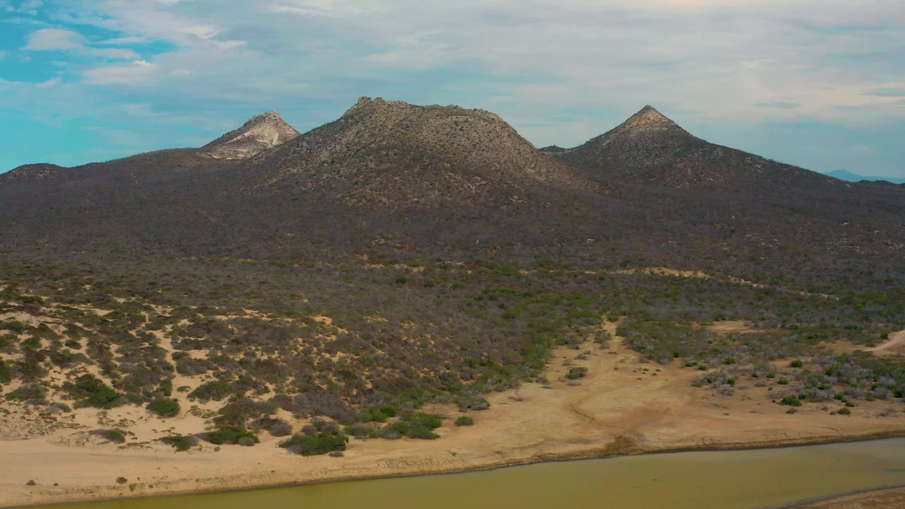 Laguna Beach, Baja California Sur aerial view with a broad sandy wash in the foreground, dark brush across the hills, three rocky peaks on the skyline, and no visible people.