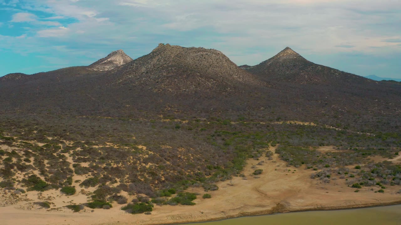 Aerial frame over Laguna Beach, Baja California Sur, with pale sand, low green shrubs, dark hillside terrain, rocky mountain peaks, a narrow waterline at the bottom edge, and no visible people.